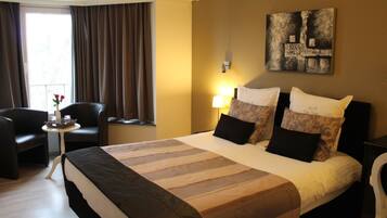Een hotelkamer met een groot bed, twee stoelen, een kleine tafel met een bloemenvaas, en een schilderij aan de muur.
