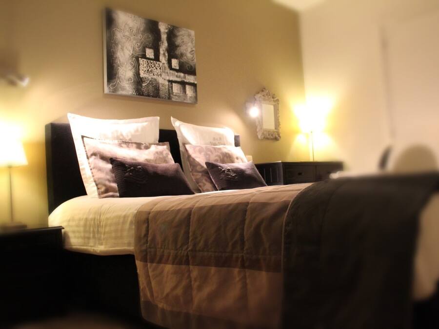 A bedroom with a bed, two bedside tables, a nightstand, a wall-mounted mirror, and a painting.