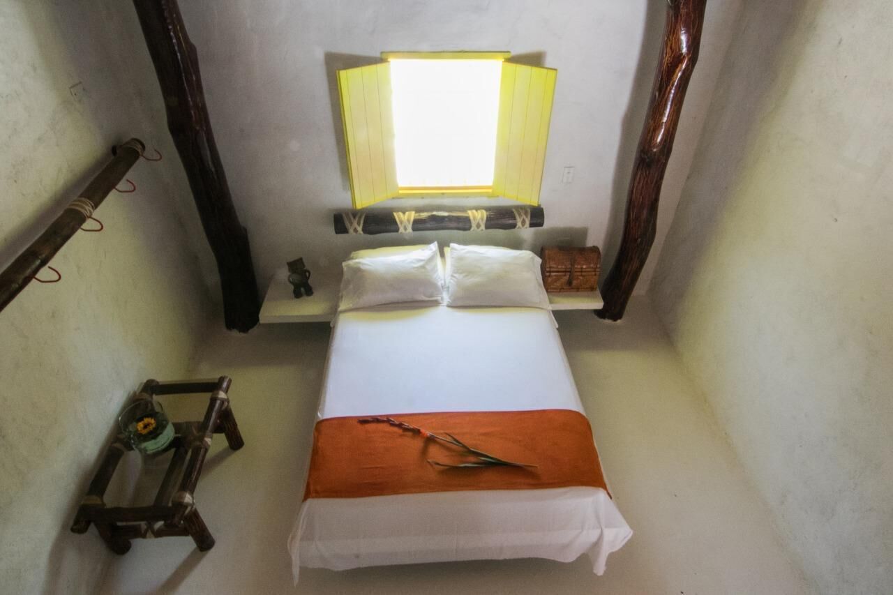 A small, narrow room with a bed, a nightstand, a lamp, and a small table with a bottle.