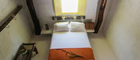 A small, narrow room with a bed, a nightstand, a lamp, and a small table with a bottle.