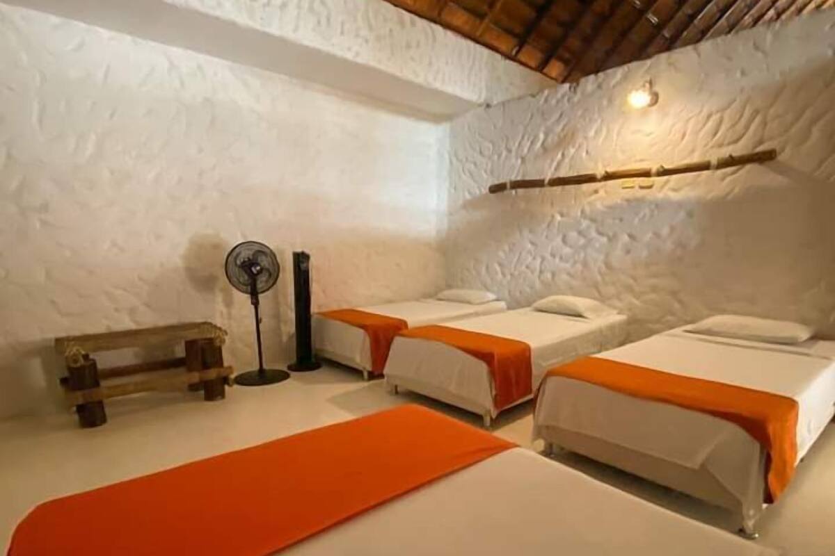 A room with three beds, each with white and orange bedding, a wooden bench, and a standing fan.
