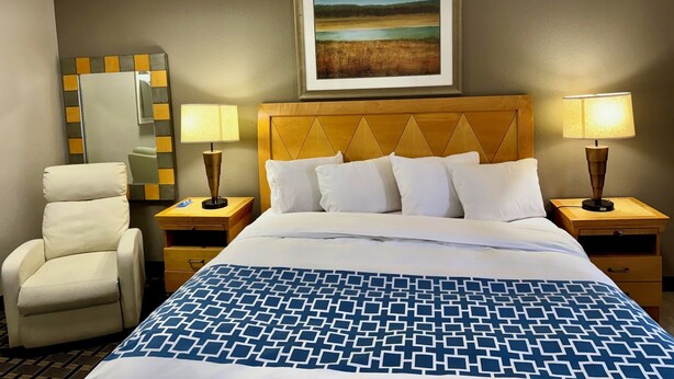 Habitación de hotel con cama, dos lámparas de noche, una silla, cómoda, espejo y un cuadro en la pared.