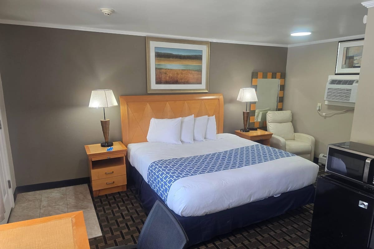 A hotel room with a bed, two bedside tables with lamps, a chair, a desk, a refrigerator, and a painting on the wall.