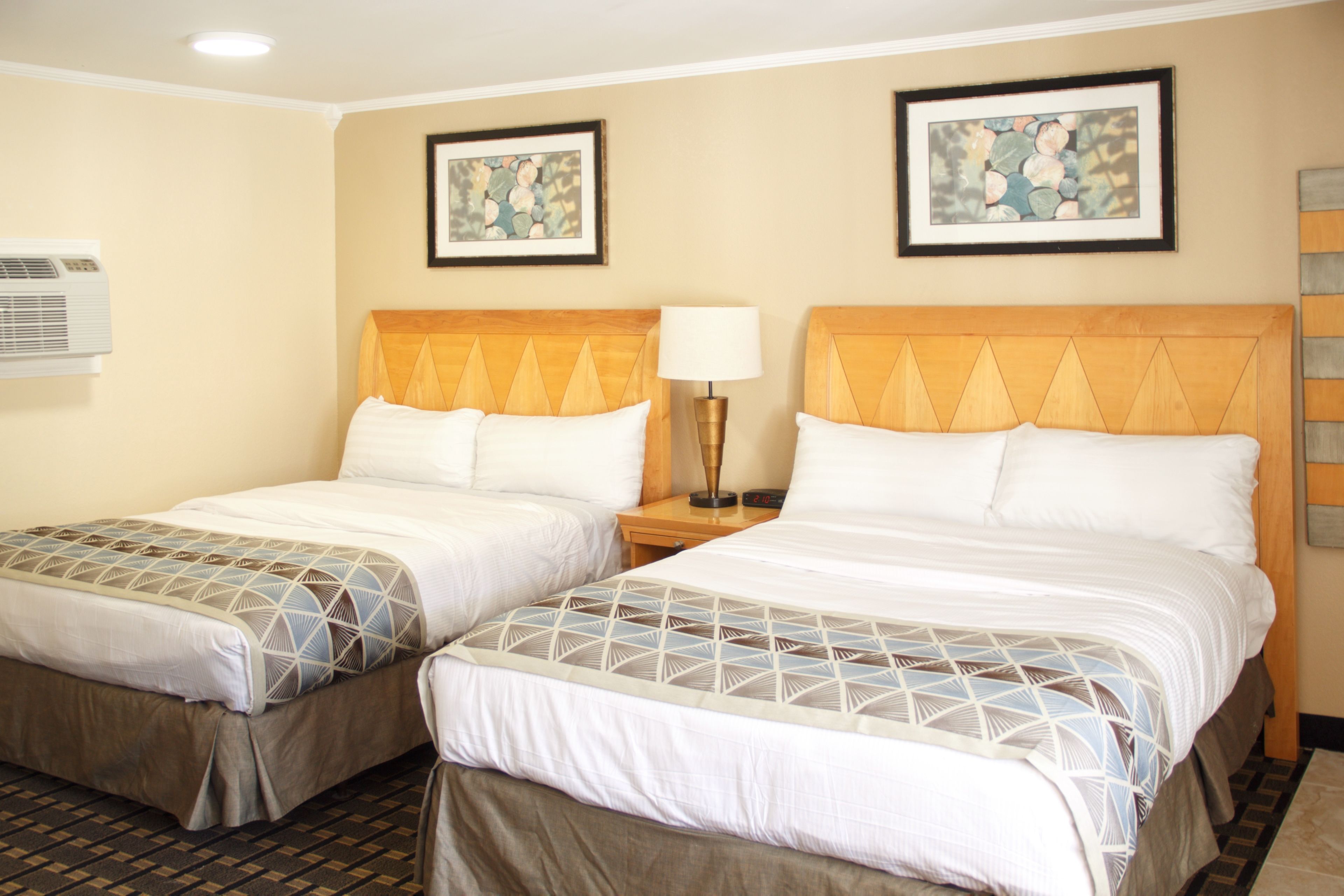 A hotel room with two beds, each with a wooden headboard and a patterned bedspread. There are framed artworks on the walls, a bedside lamp, and an air conditioning unit.