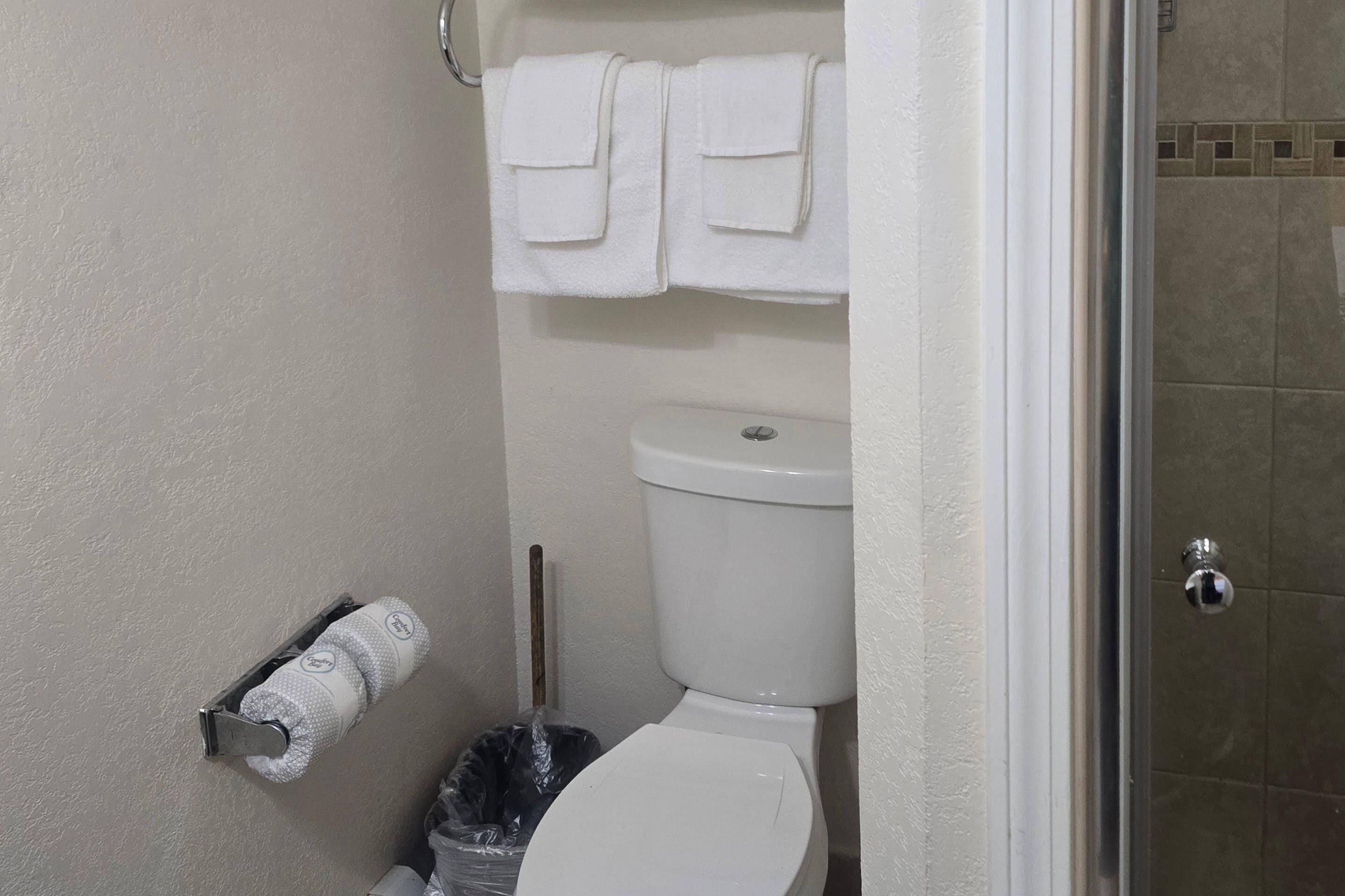 A bathroom with a toilet, a towel rack with two towels, a toilet paper holder, and a trash bin.