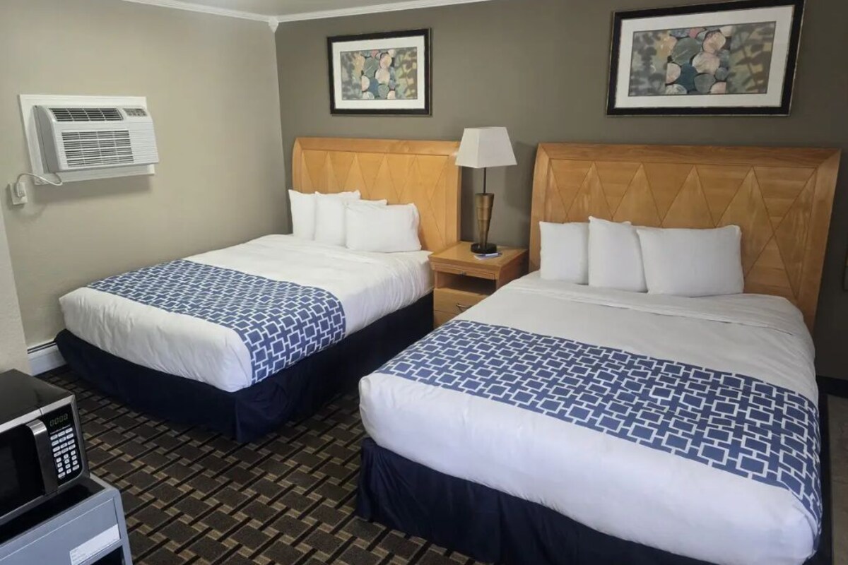 A hotel room with two beds, a nightstand, a lamp, an air conditioning unit, and a microwave.