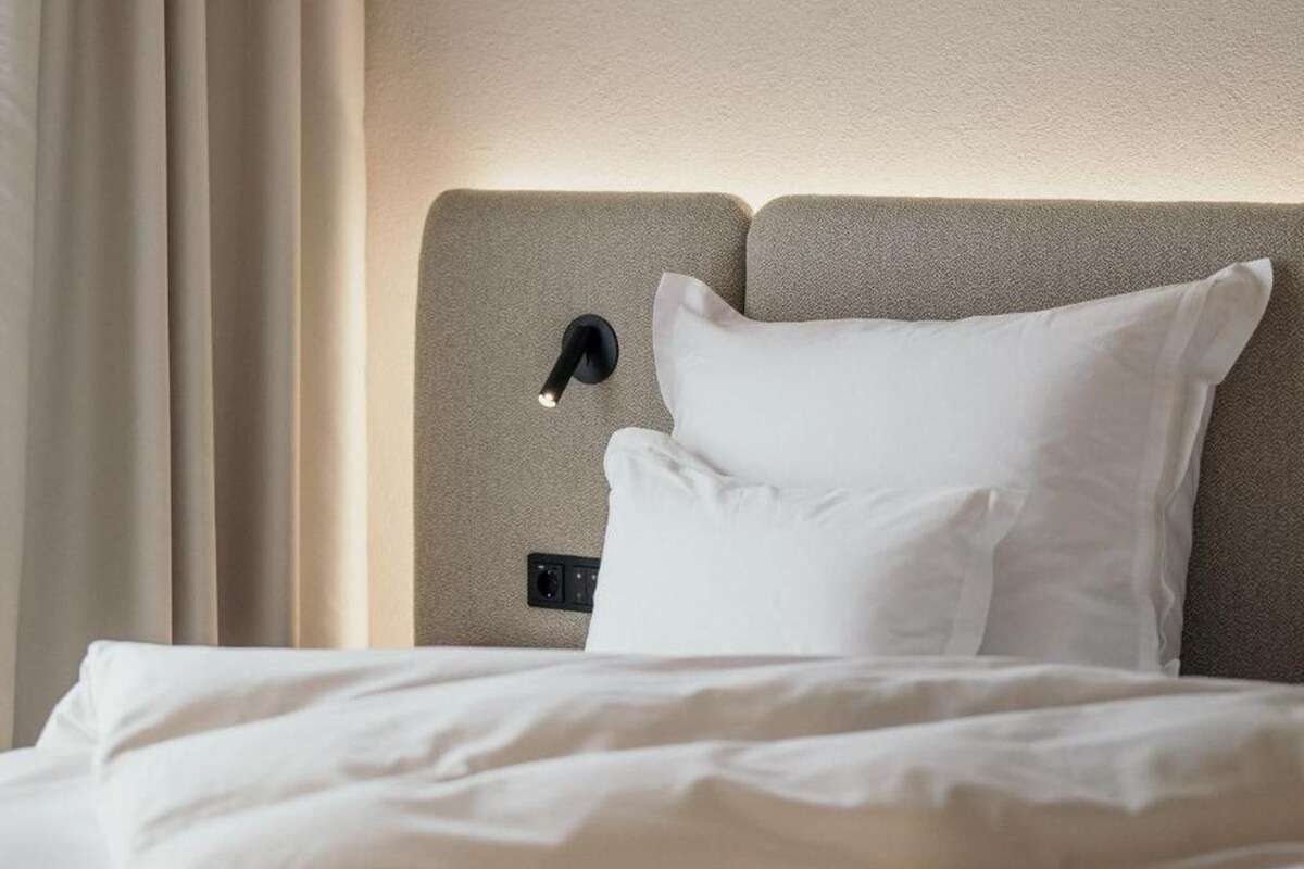 A hotel room with a bed, a headboard, a wall-mounted light, and a window with curtains.