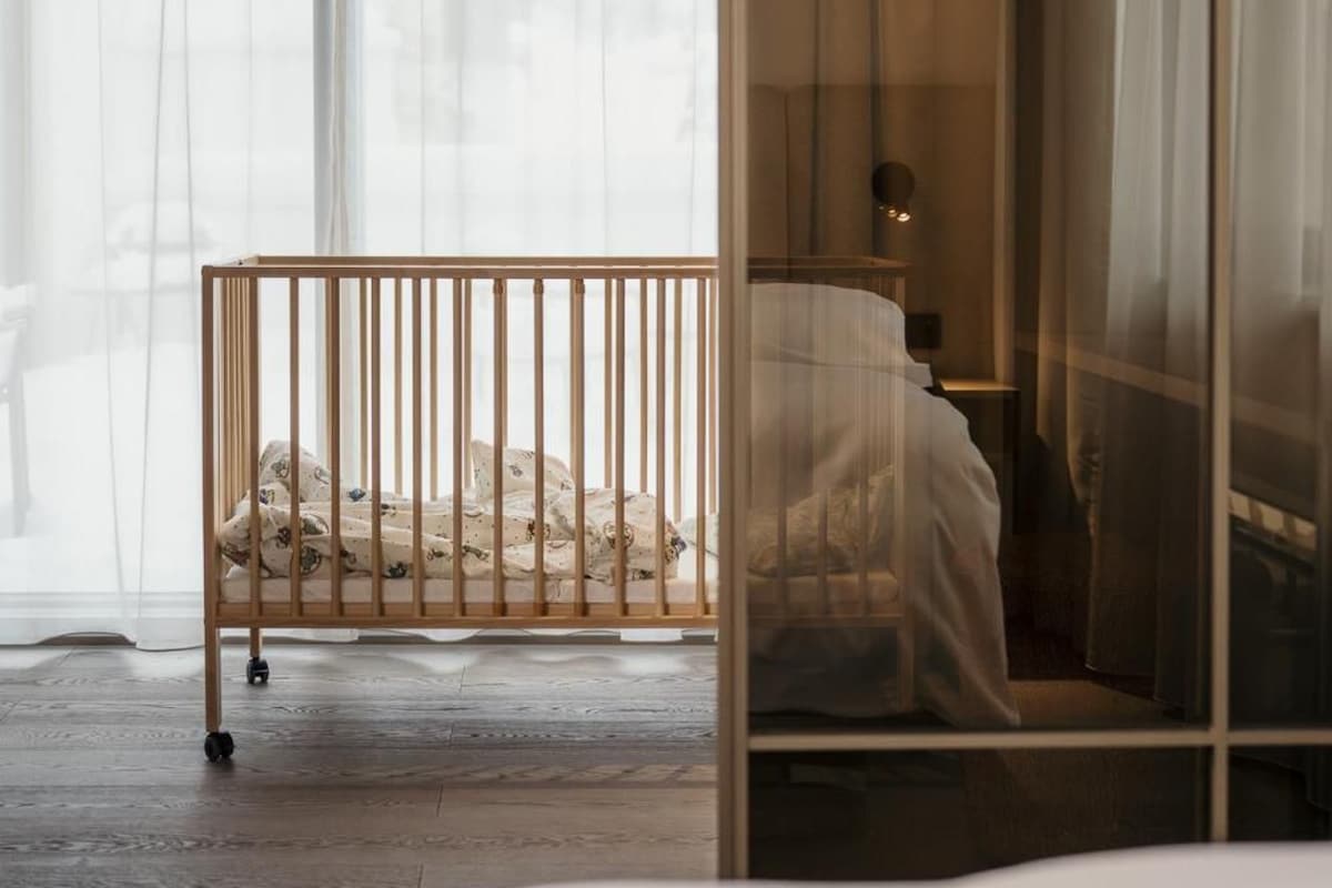 A room with a wooden crib, a bed with a white bedspread, and sheer curtains.