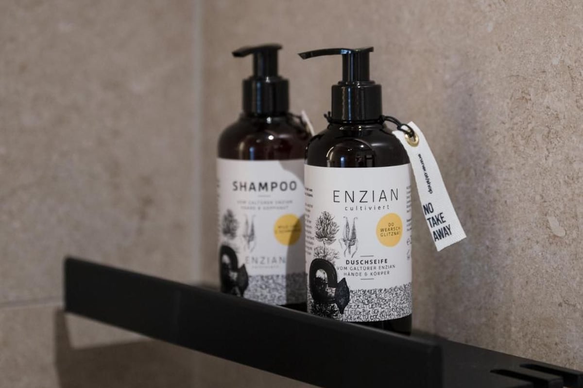 Dos botellas de champú y acondicionador Enzian sobre un estante negro frente a una pared con textura.