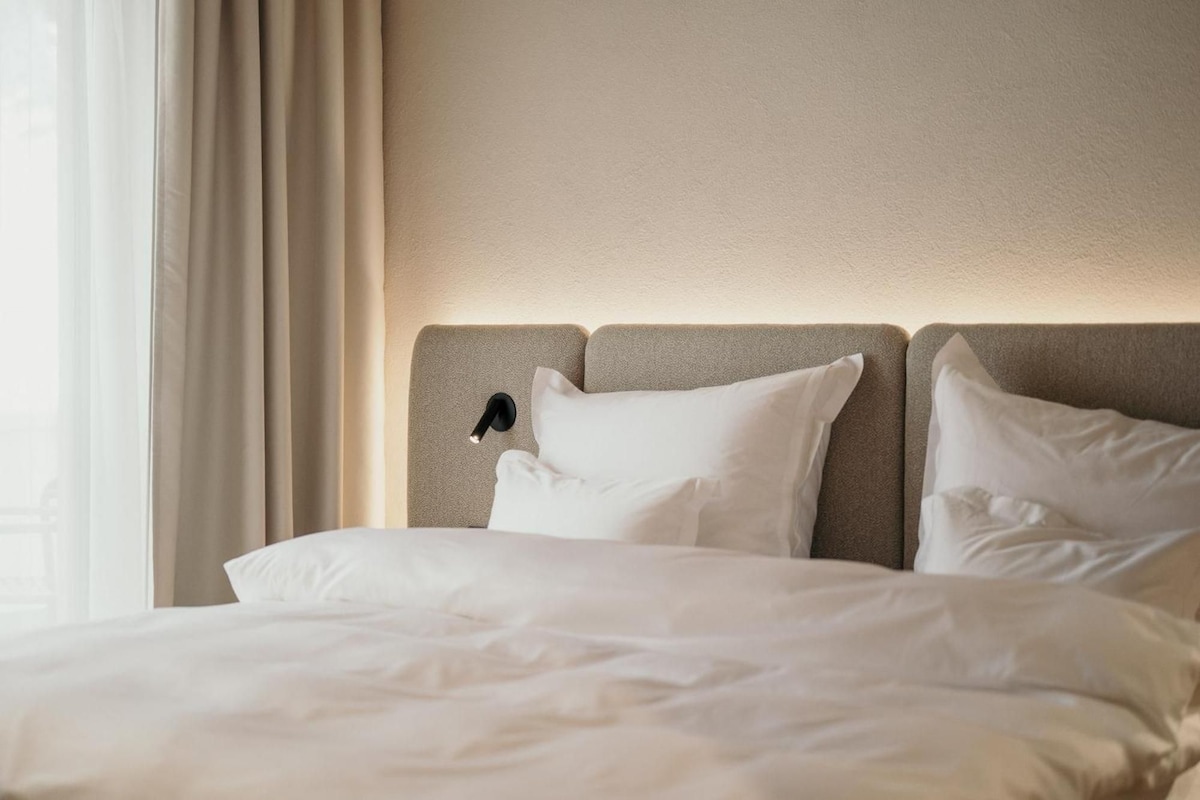 Una habitación de hotel con cama, ropa de cama blanca, cabecero y ventana con cortinas.