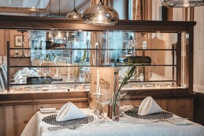 A restaurant interior with a wooden paneled wall, hanging pendant lights, and a glass display case with wine bottles and glasses.