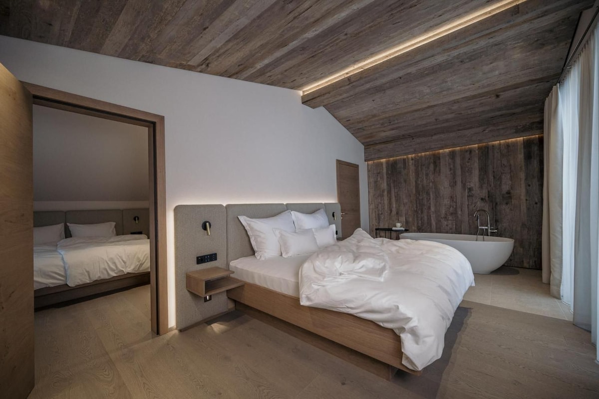 A modern bedroom with a large bed, bedside tables, a built-in bench, a large window, and a built-in bathtub.