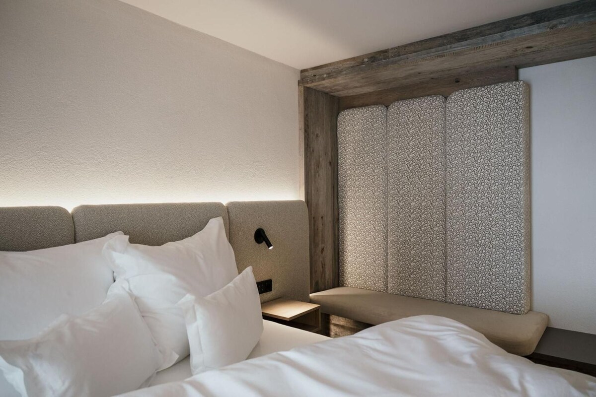 A hotel room with a bed, a nightstand, a bench, and a patterned curtain.