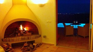 Una chimenea encendida, un ventanal de vidrio que da a un área de estar exterior con una mesa y sillas, y vistas al cielo nocturno.