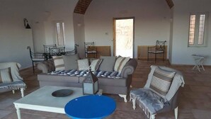 Amplia sala de estar con un conjunto de sofás, una mesa de centro y un tapete azul. Hay dos sillones y una mesita. La habitación tiene un techo alto con vigas a la vista y un ventanal que proporciona luz natural.