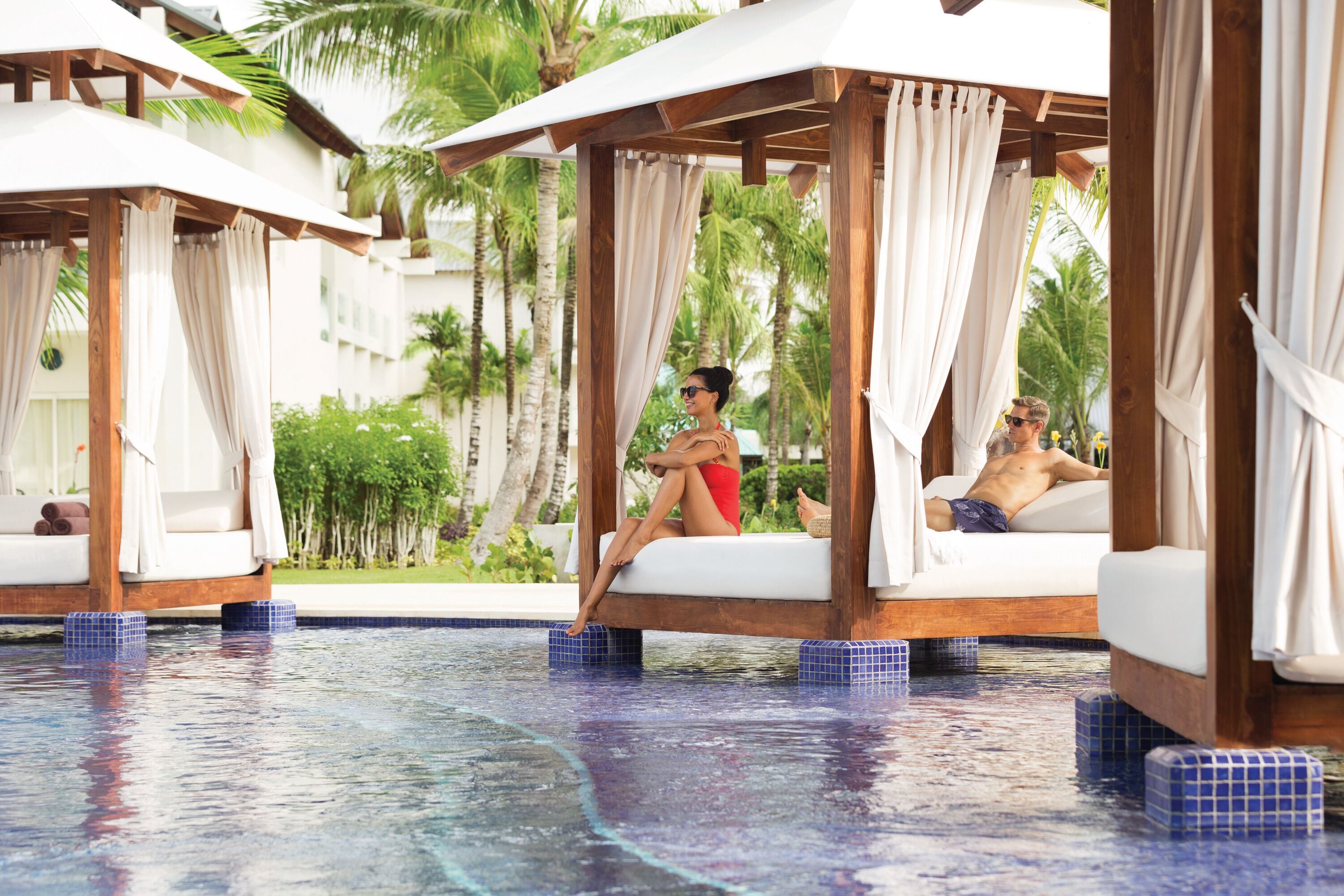 A pool area with wooden cabanas and white cushions, surrounded by palm trees.