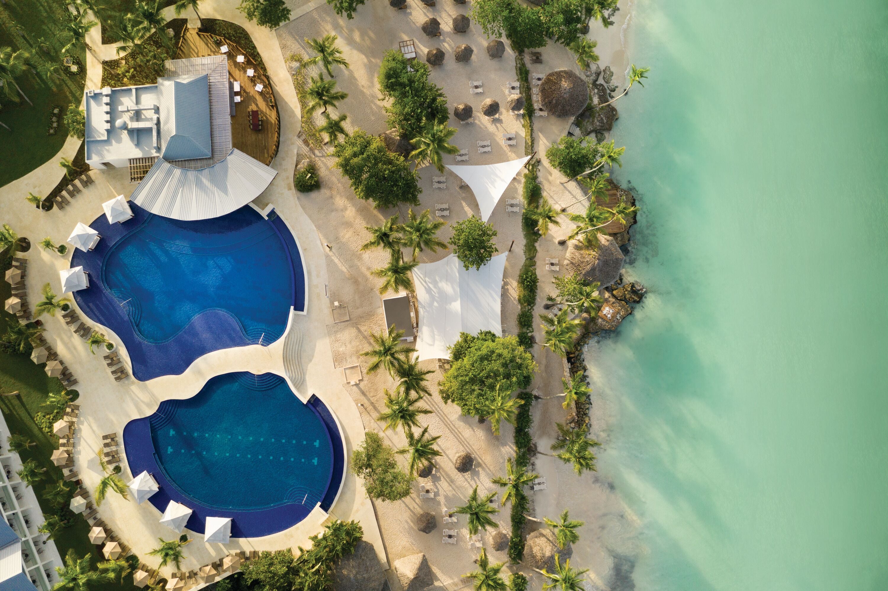 An aerial view of a resort with two large swimming pools, surrounded by palm trees and beach chairs.