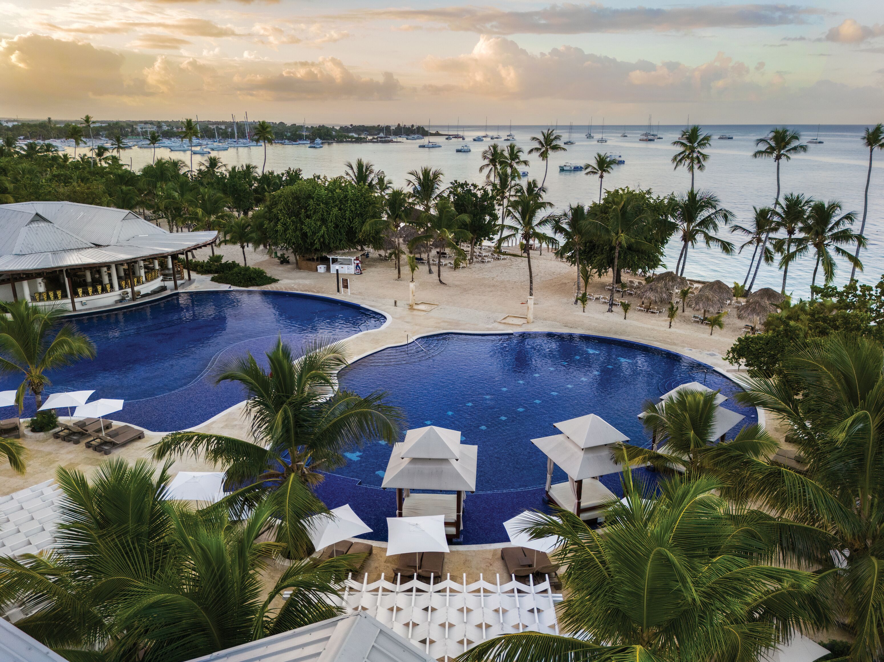 A resort with two large swimming pools, a beach area with umbrellas, and a view of the ocean with boats.