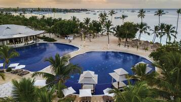 A resort with two large swimming pools, a beach area with umbrellas, and a view of the ocean with boats.