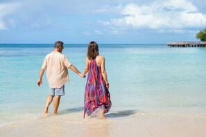 Una pareja caminando de la mano por una playa con agua cristalina y un muelle al fondo.