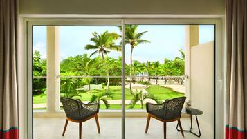 A balcony with two wicker chairs and a small table, overlooking a lush garden with palm trees and a pool.