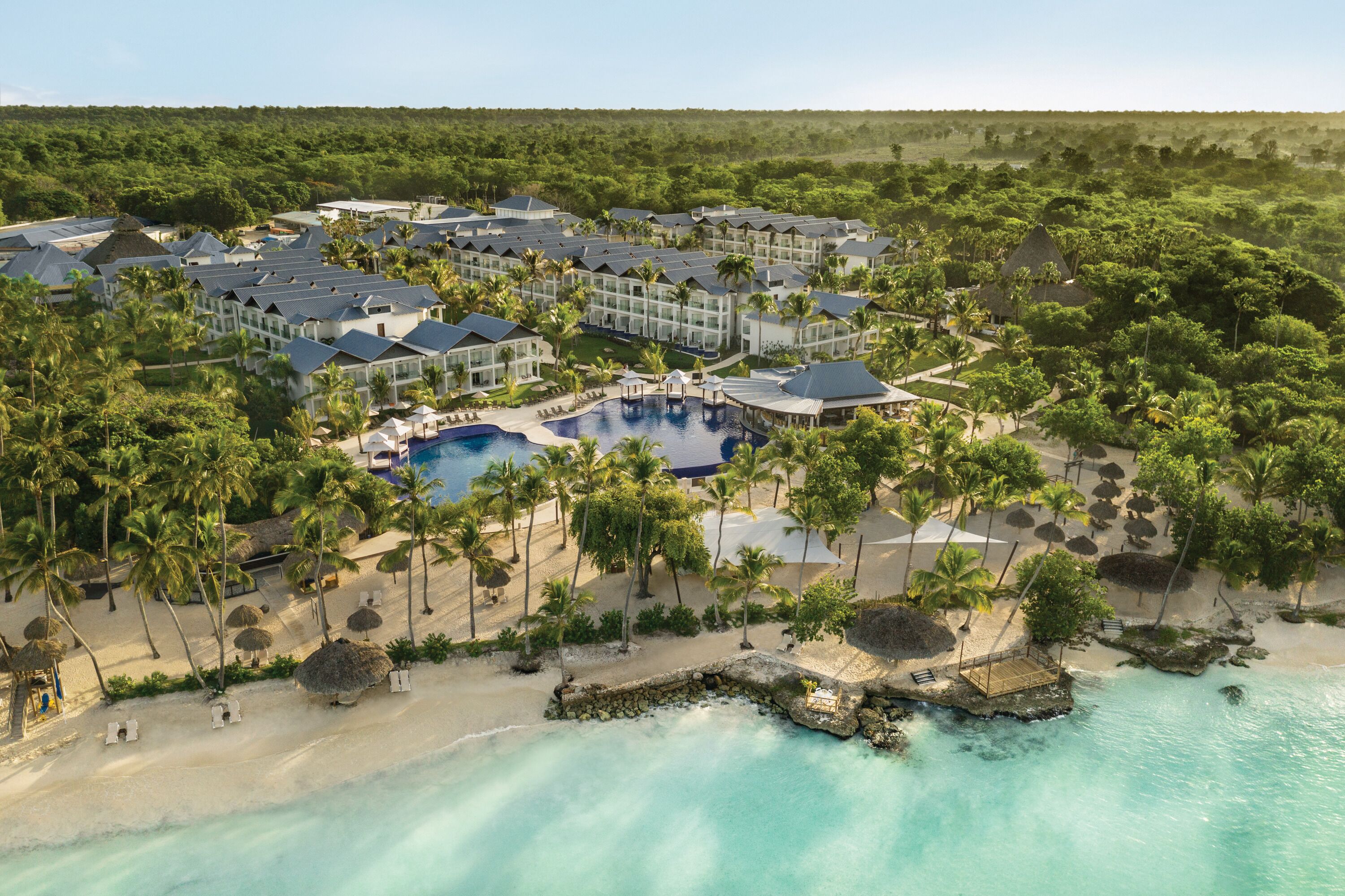 Vista aerea di un resort con piscina centrale, circondato da palme e spiaggia.