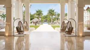 A hotel lobby with a central pathway leading to a pool area, surrounded by palm trees and thatched-roof structures. - Secrets La Romana Resort & Spa  - Adults Only - All Inclusive (San Rafael del Yuma)