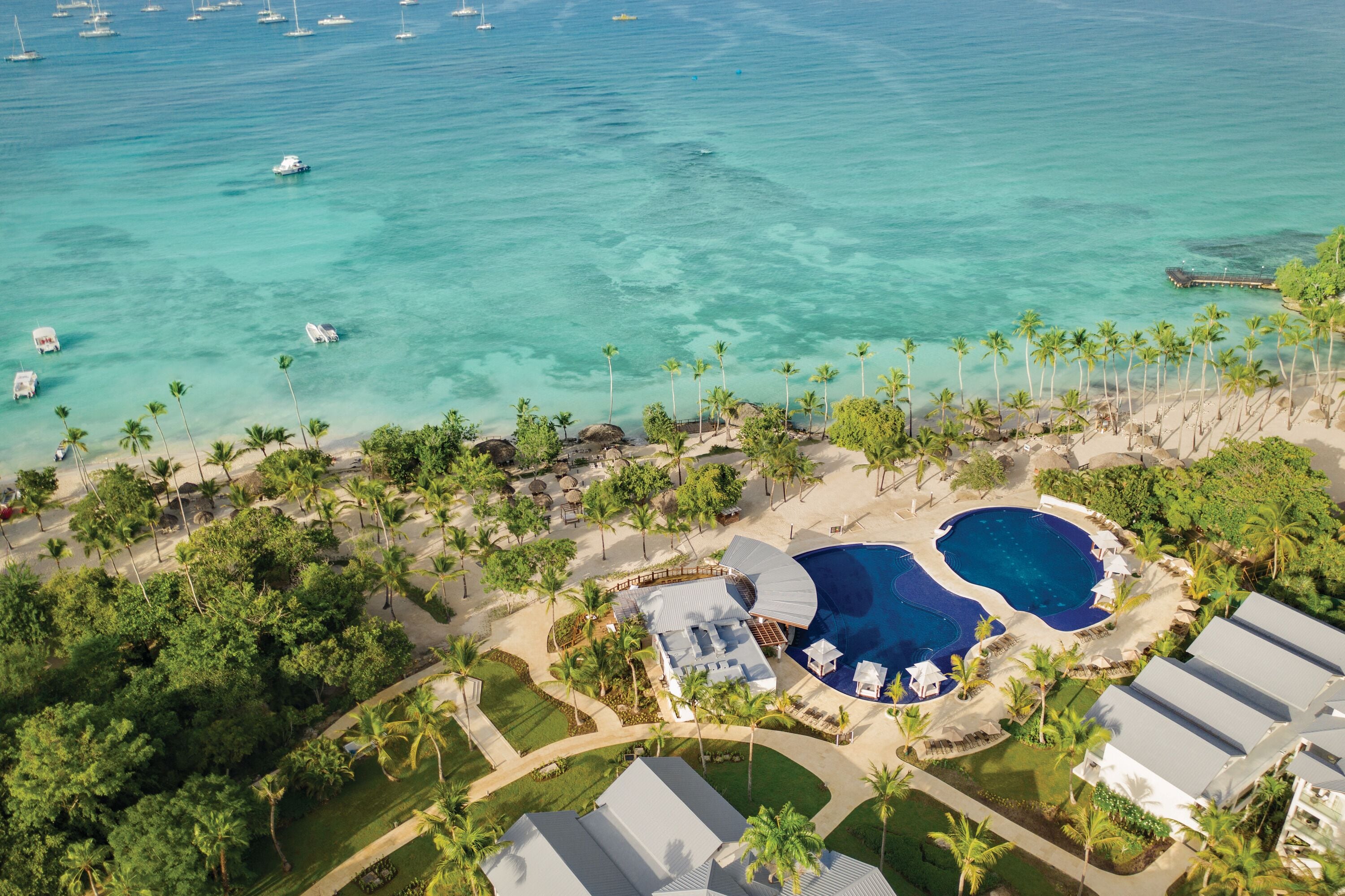 An aerial view of a resort with two large swimming pools, a beach area with palm trees, and a marina with boats.