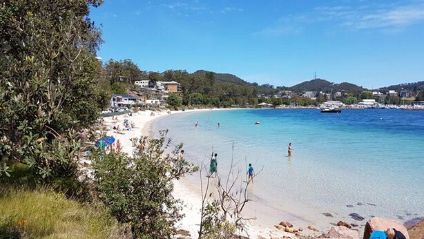 Ein Strand mit klarem Wasser, badenden Personen und Gebäuden im Hintergrund.