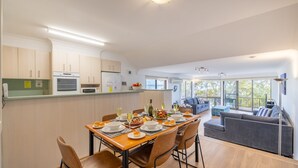 Eine moderne Küche mit eingebauten Geräten, ein Essbereich mit Holztisch und -stühlen sowie ein Wohnbereich mit einer Couch und großen Fenstern.