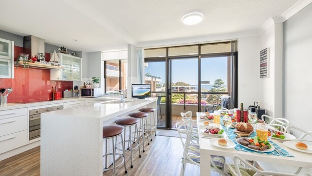 A modern kitchen with a large island, bar stools, and a dining area with a table set for a meal. - The Commodore, 23,9-11 Donald Street - Delightful unit with gorgeous water views, air con and wi-fi (Nelson Bay)