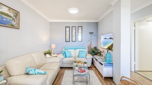 A modern living room with a sofa, a coffee table, and a TV. There are three framed pictures on the wall, a lamp, and a plant on the side table. - The Commodore, 23,9-11 Donald Street - Delightful unit with gorgeous water views, air con and wi-fi (Nelson Bay)