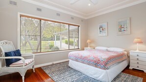 Ein Schlafzimmer mit großem Fenster, einem Bett mit Blumenmuster, einem weißen Sessel mit Kissen, einem Nachttisch mit Lampe und zwei gerahmten Bildern an der Wand.