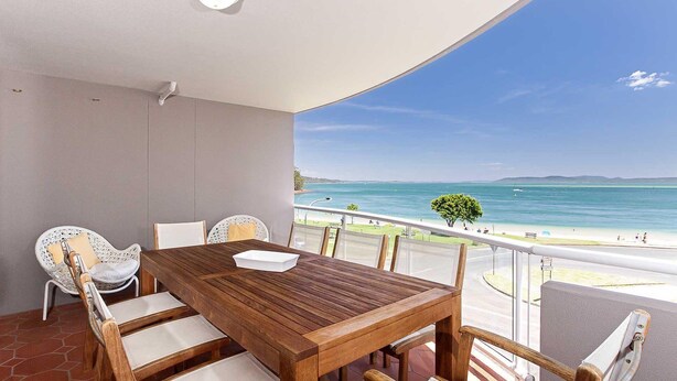 A balcony with a wooden table and chairs, overlooking a beach with clear blue water and a distant shoreline. - Florentine, 12/11 Columbia Close - WIFI, Aircon, spectacular views (Nelson Bay)