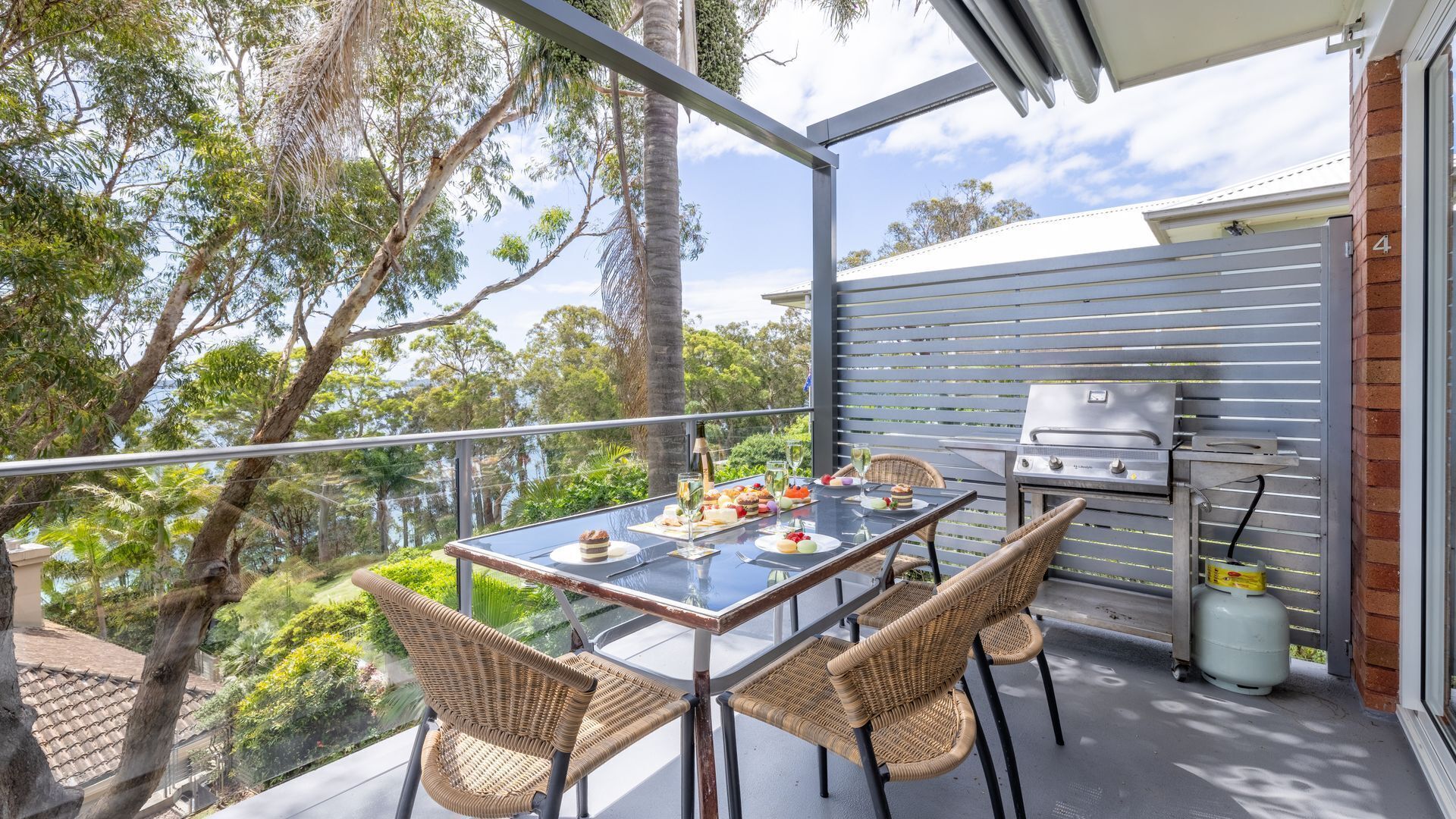 An outdoor dining area with a table set for four, surrounded by wicker chairs, overlooking a lush garden and a brick building.
