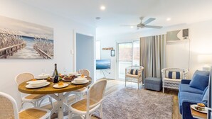 A modern living room with a dining area, wicker furniture, a flat-screen TV, and a large window with curtains. - Hibiscus Court, 2/9 Government Road - fantastic air conditioned 3 bedroom unit (Shoal Bay)