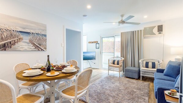 A modern living room with a dining area, wicker furniture, a flat-screen TV, and a large window with curtains.
