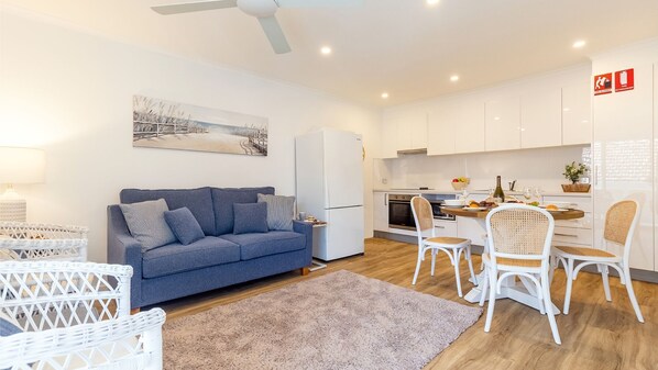 A modern living room with a sofa, a dining table with chairs, a kitchen area with cabinets and appliances, and a ceiling fan. - Hibiscus Court, 2/9 Government Road - fantastic air conditioned 3 bedroom unit (Shoal Bay)