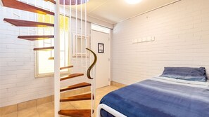 A bedroom with a bed, a blue blanket, a wooden staircase, and a white brick wall. - Hibiscus Court, 2/9 Government Road - fantastic air conditioned 3 bedroom unit (Shoal Bay)