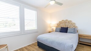 A bedroom with a bed, a ceiling fan, a bedside table with a lamp, and a window with blinds. - Hibiscus Court, 2/9 Government Road - fantastic air conditioned 3 bedroom unit (Shoal Bay)