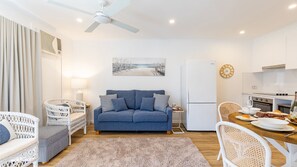 A modern living room with a blue sofa, white wicker furniture, a wooden dining table with plates and glasses, and a kitchen area with a white refrigerator and stove.