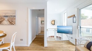 A modern living room with a wooden floor, a dining area with wicker chairs, a TV mounted on the wall, and a view of the sea through large windows. - Hibiscus Court, 2/9 Government Road - fantastic air conditioned 3 bedroom unit (Shoal Bay)