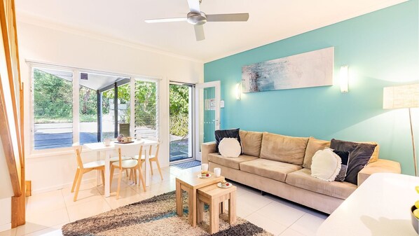 Una sala de estar con un sofá color beige, una mesa de centro y un comedor con una mesa y sillas. Hay un ventilador de techo, un cuadro en la pared y una ventana con vista a la vegetación exterior.