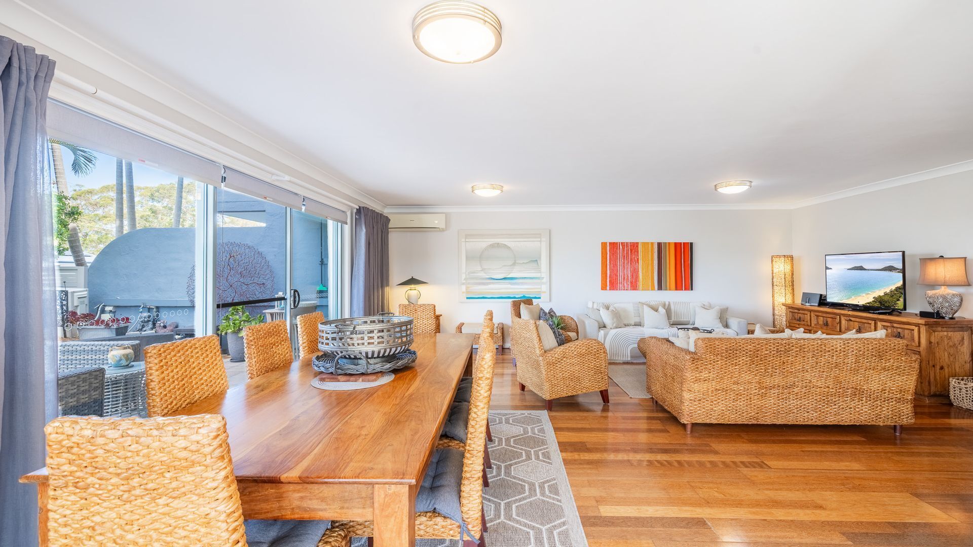 A spacious living area with a wooden dining table, wicker furniture, and a large window offering a view of the outdoors.
