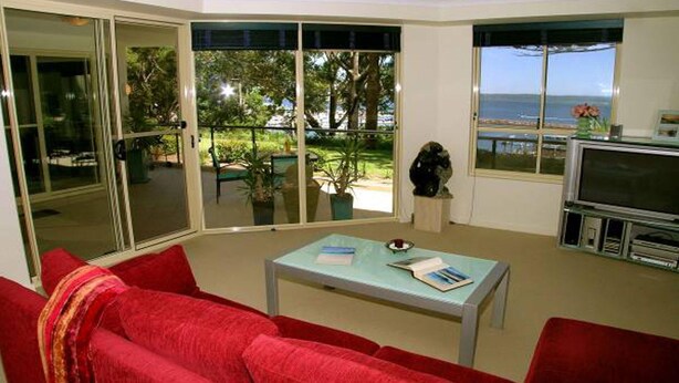 Un salón con un sofá rojo, una mesa con libros, un televisor y ventanas grandes con vistas al mar.