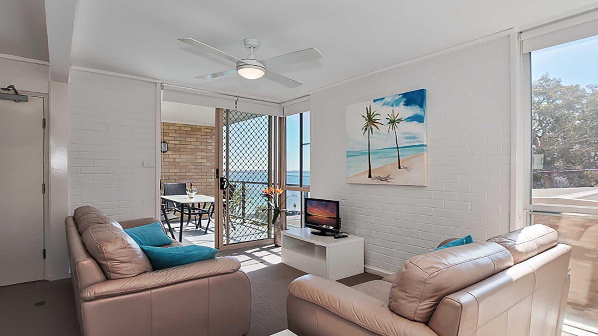 A modern living room with a TV, leather sofas, and a balcony with a view of the sea.