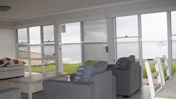 En rummelig stue med store vinduer, der giver udsigt over en sø og både. Der er en grå sofa, en hvid lænestol og et lille bord.