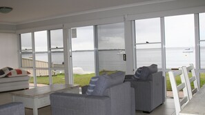 A spacious room with large windows offering a view of a lake and boats. There is a grey sofa, a white armchair, and a small table. - The Clippers, 1/131 Soldiers Point Road - fabulous waterfront unit (Soldiers Point)