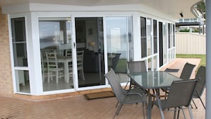 En overdækket terrasse med et glasindhegnet rum, et bord med stole og udsigt til baghaven.