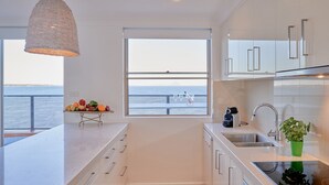 Una cocina moderna con un amplio ventanal con vista al mar, una lámpara colgante y una pequeña mesa con frutas y vasos.